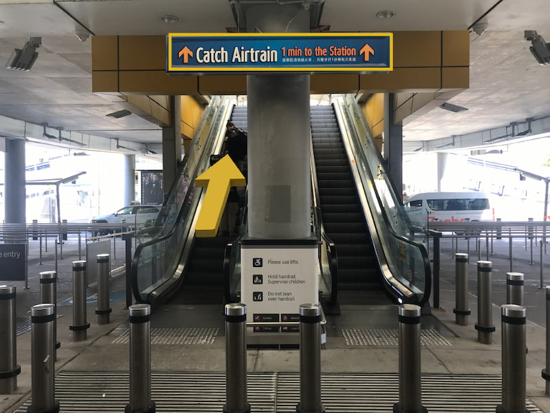 Escalator underneath airport skywalk, blue sign that reads Catch Airtrain with arrows pointing up.